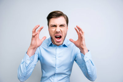 portrait of very angry, annoyed, wild man shouting, yelling, holding hands near face, standing over grey background