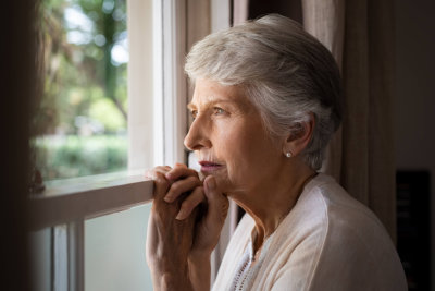 depressed senior woman at home feeling sad. Elderly woman looks sadly outside the window. Depressed lonely lady standing alone and looking through the window.